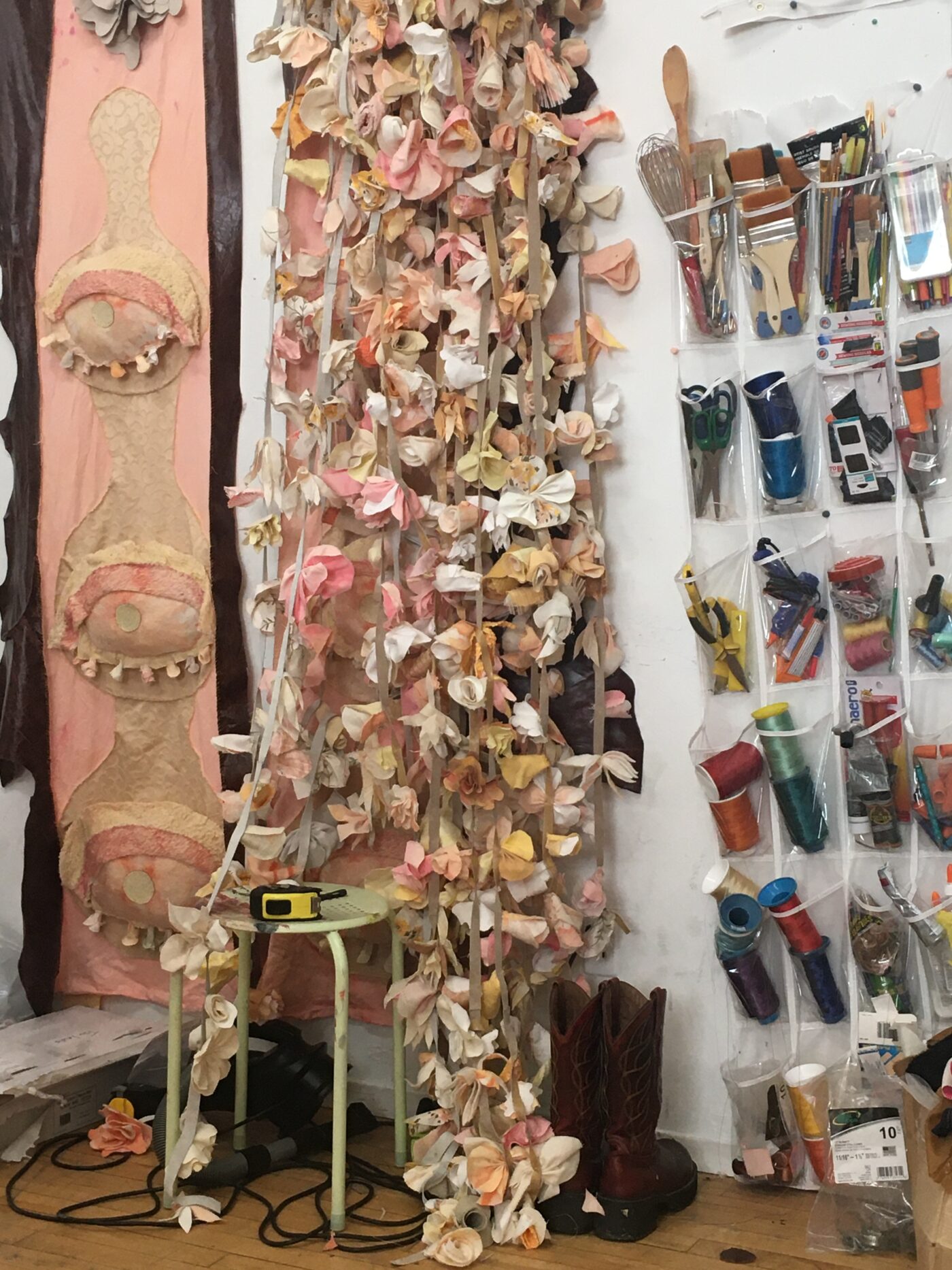 Image shows a wall. Hanging on the left of the image is an artwork – a banner hangs vertically. The banner is pink-flesh coloured and has applique eyelid textile sculptures along the length. In the centre are dozens upon dozens of textile flower strings. They vary in colour from pinks to oranges to whites. On the right on the image are plastic organizational pouches containing the tools of an artist: paintbrushes, scissors, string and various other tools.