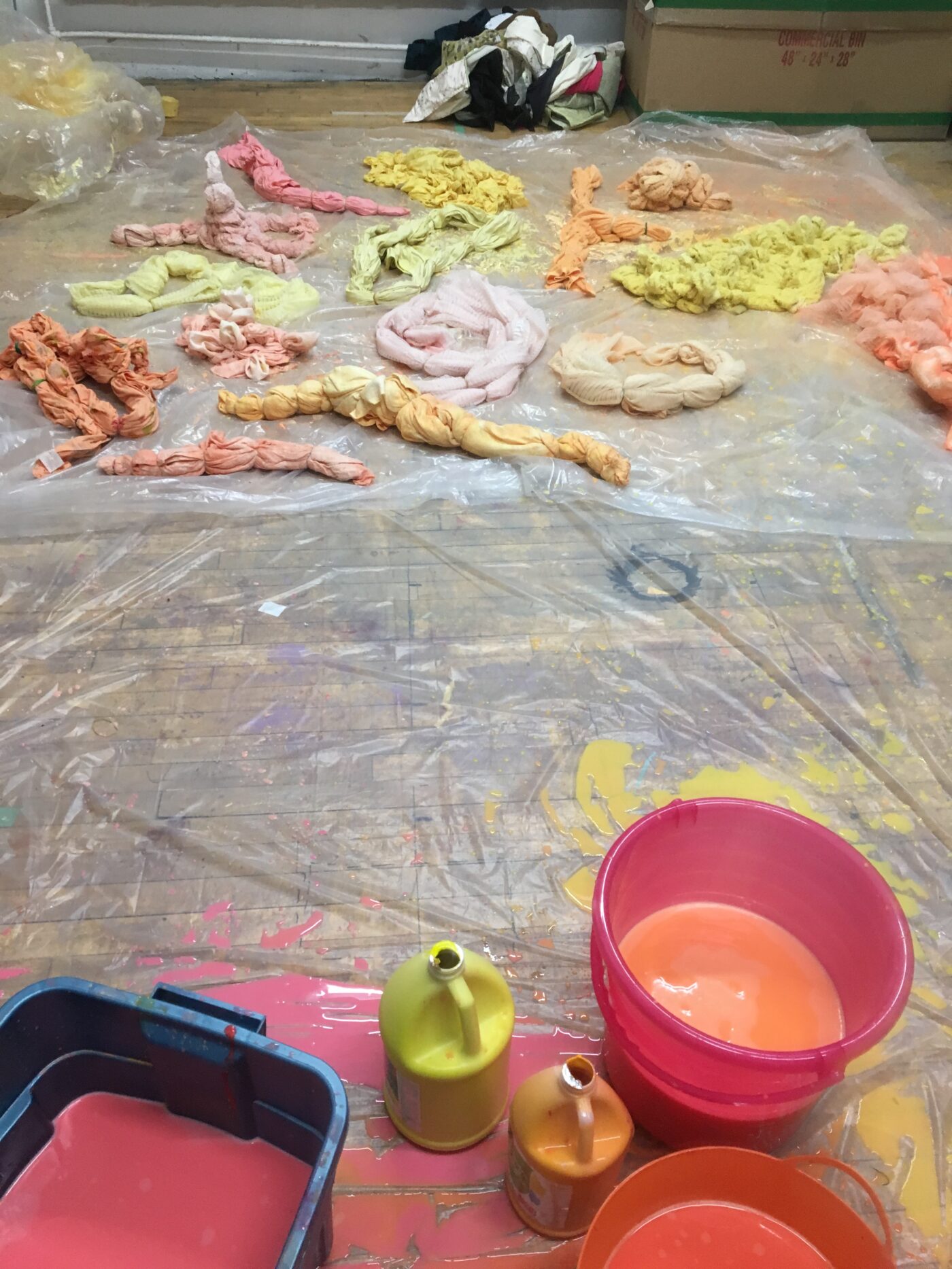 An image of Tau Lewis' process – in the foreground on the image are buckets of paint resting on a plastic sheet on the floor. In the background are textile pieces, likely dipped into the paints, coiled, and left to dry.