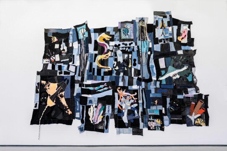 Image shows Tau Lewis' piece "The coral reef preservation society". A large, uneven quilt made from dark fabrics. Across the fabric are applique sea creatures, created from various swathes of colourful fabric. There is a sea horse, jellyfish, a sting ray, a turtle, a hammerhead shark, coral, and other small fish. 