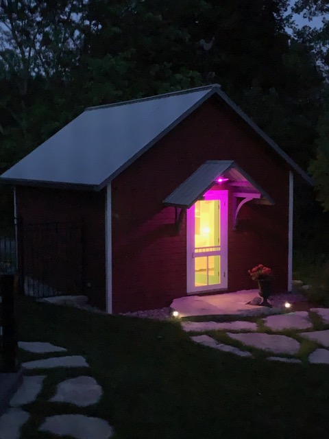 The image shows a one-storey structure with a pitched roof and red siding. There is a door in the middle of the narrow side of the structure that is lit from the inside. The structure has trees in the background and a stone path leading to it