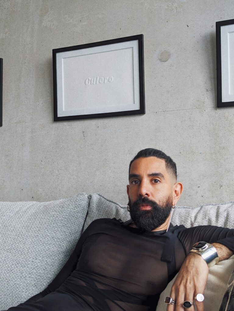 Armando Perla sitting on a white couch against a white wall. They are wearing a black sheer top with solid black crisscrossing ties. They are wearing a silver watch and rings while leaning against the arm of the couch.